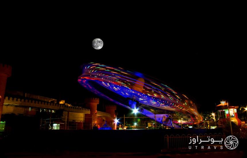 پارک ارم تهران در تاریکی شب؛ یک وسیله بازی درحال چرخش و ماه بالای سر آن